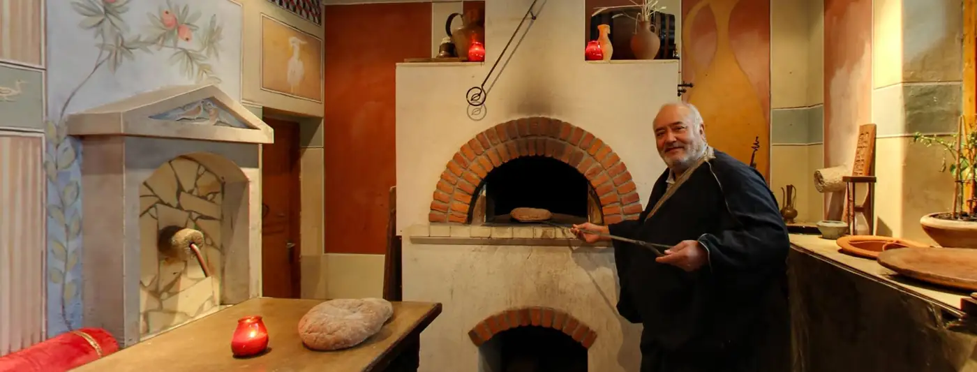 Man in donker gewaad bij een stenen oven in een Romeins interieur met een fontein op de achtergrond
